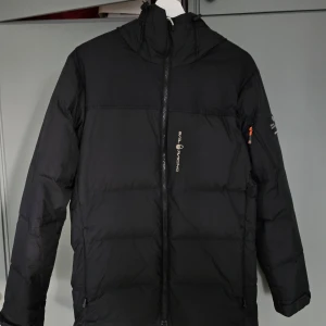 Svart dunjacka från Sail Racing S - Svart dunjacka från Sail Racing med huva och dragkedja framtill. Jackan är vadderad och har logga på bröstet samt på ryggen. Tillverkad i nylon och fylld med 70% dun och 30% fjäder. Perfekt för kalla dagar när du vill hålla värmen.