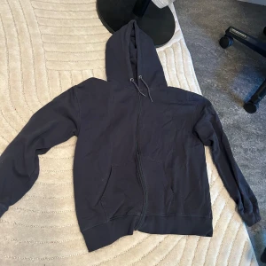 Svart hoodie med dragkedja - Svart hoodie med luva och snörning, hel dragkedja framtill och två stora fickor. Perfekt för chill dagar eller när du vill ha något bekvämt och enkelt. Långärmad och i mjukt bomullsmaterial som känns skönt mot huden.