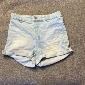 Ljusblå jeansshorts från Divided i storlek 36 - Snygga ljusblå jeansshorts från Dividedi storlek 36 med klassisk femficksdesign och uppvikta benslut. Shortsen har normal passform, bälteshällor och knappstängning fram. Perfekta för varma dagar och chill häng med kompisarna.
