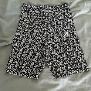 Svarta och vita mönstrade shorts Adidas - Snygga svarta shorts från Adidas med vitt grafiskt mönster över hela tyget och diskret logga på ena benet. De har elastisk midja och är tillverkade i ett lätt och bekvämt material, perfekta för träning eller chill dagar.