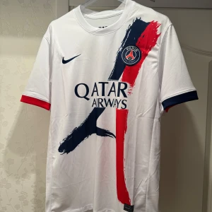 PSG vit matchtröja Nike - Snygg PSG fotbollströja från Nike i vit polyester med coola penseldrag i marinblått och rött på framsidan. Paris Saint-Germain-logga på bröstet och Qatar Airways-tryck. Kortärmad med kontrastfärgade muddar i blått och rött. Perfekt för dig som älskar fotboll och stil.