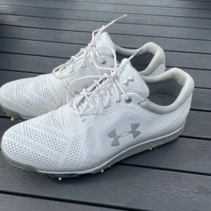 Vita golfskor från Under Armour - Säljer ett par vita golfskor från Under Armour med snörning och perforerat skinn för extra andningsförmåga. Skorna har tydliga dobbar undertill för bra grepp på golfbanan och en sportig look med logga på sidan. Perfekta för dig som vill ha stil och funktion på greenen. Skorna är sparsamt använda och säljer dem då de inte riktigt passade min fötter. Nypris 1250 kr villl bara få sålt dem nu