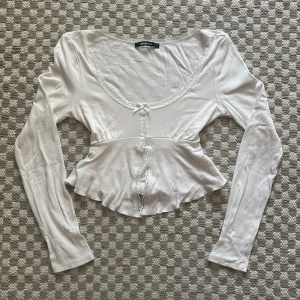 Vit croppad topp med spets från Gina Tricot - Supersöt vit croppad topp från Gina Tricot med långa ärmar, spetsdetaljer längs halsringningen och framtill, samt en liten rosett. Toppen har en U-ringad form och är tillverkad i mjuk bomullsmix. Perfekt att styla med jeans eller kjol för en trendig look.