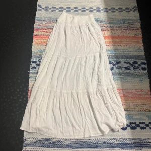 Vit långkjol från 157 med resår - Luftig vit långkjol från 157 med bred smockresår i midjan. Kjolen har flera volanger och är tillverkad i ett mjukt, tunt tyg som ger en flowy känsla. Perfekt för varma dagar och enkel att styla med crop tops eller linnen.