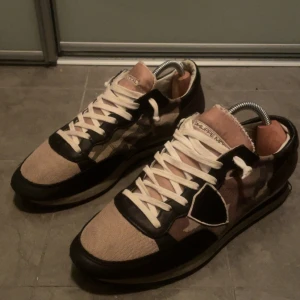 Philip model sneakers camouflage - Snygga Philip model sneakers med camouflagemönster i grönt, beige och brunt. Skorna har svarta skinnpaneler, beige textil på tå och sidor samt vita snören. Ikonisk design med platt sula och rund tå, perfekt för dig som gillar streetstyle och exklusiva märken. Strl 42