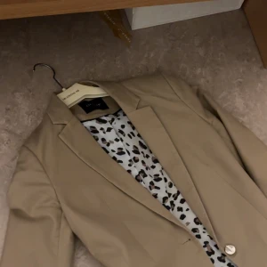 Beige kavaj från Only - Snygg beige kavaj från Only med klassisk krage och knapp framtill. Insidan har ett coolt leopardmönstrat foder som ger extra edge. Perfekt för dig som vill ha en stilren look med en twist.