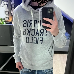 Grå hoodie med tryck och huva - Säljer en ljusgrå hoodie med svart texttryck på bröstet. Tröjan har huva med vita snören och en stor magficka framtill. Perfekt för dig som gillar en avslappnad och sportig stil.
