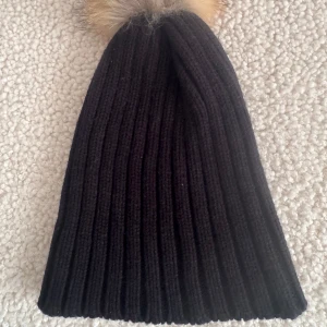 Svart ribbad mössa med pälsboll - Säljer en svart, ribbstickad mössa med stor fluffig pälsboll på toppen. Mössan är i stickad material och har en bred uppvikt kant. Perfekt för dig som vill ha en mysig och snygg accessoar till vintern. Pälsbollen är i beige och bruna toner.