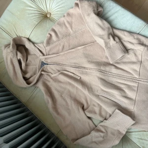 Beige stickad hoodie från Cashmere - Mysig beige stickad hoodie från Cashmere med dragkedja framtill, huva med snörning och två fickor. Tröjan har ribbade muddar och är långärmad, perfekt för lager-på-lager och chill dagar.