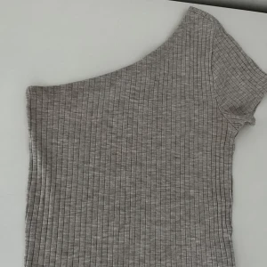 Grå ribbad oneshoulder topp - Snygg grå ribbad topp med oneshoulder-design och kort ärm. Perfekt för dig som gillar en stilren och modern look. Mjuk och stretchig, passar till många outfits.