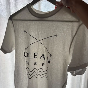 Vit t-shirt Ocean Vibes Tribute S - Vit t-shirt från Tribute i storlek S med svart tryck 'Ocean Vibes', två korsade pilar och vågmönster. Klassisk rund hals och korta ärmar. Mjuk och lätt bomullskvalitet, perfekt för chill dagar. Snyggt grafiskt print framtill.
