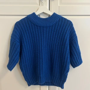 Blå stickad tröja med kort ärm - Säljer en chunky blå stickad tröja med korta ärmar och bred ribbad krage. Perfekt för lager-på-lager och ger en cool vibe till din outfit. Tröjan har en loose fit och är supermjuk och skön att bära.