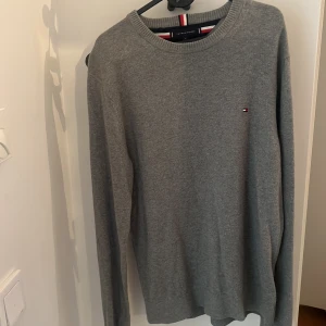 Grå stickad tröja Tommy Hilfiger M - Snygg grå stickad tröja från Tommy Hilfiger i pima bomullsblandning med kashmir. Klassisk rund halsringning och liten broderad logga på bröstet. Perfekt för dig som gillar stilrena och bekväma plagg.