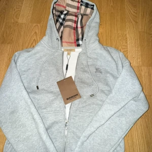 Grå hoodie från Burberry med dragkedja -  Snygg grå  hoodie från Burberry med klassiskt rutigt foder i huvan och broderad logga på bröstet. Tröjan har dragkedja, snörning i huvan och är tillverkad i mjuk bomull. Perfekt för en chill och stilren look.