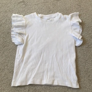 Vit ribbad topp med volangärmar - Superfin vit ribbad topp med korta volangärmar. Perfekt för dig som gillar en söt och stilren look. Toppen har rund halsringning och är tillverkad i mjuk bomull som känns skön mot huden. Pris kan diskuteras 