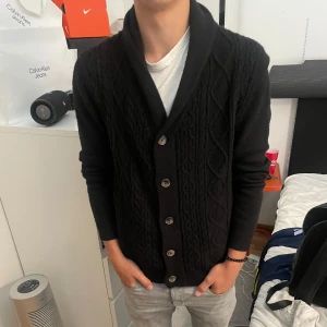 Svart stickad kofta från Threadbare - Svart stickad kofta från Threadbare med snyggt kabelstickat mönster och bred sjalkrage. Koftan har långa ärmar och stängs med fem knappar framtill. Perfekt för lager-på-lager och chill höst- och vinterstil.