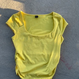 Gul topp med rynkade sidor  - Superfin gul topp med rundad, djup urringning och korta ärmar. Toppen har snygga rynkade detaljer i sidorna som ger en cool twist. Perfekt för dig som gillar färg och vill sticka ut lite extra i sommar.