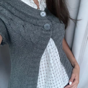 Grå stickad kofta med flätmönster - Säljer en grå stickad kofta med korta ärmar och snyggt flätmönster över bröstet. Koftan har två stora knappar framtill och bred ribbad kant. Vet ej vad koftan kommer ifrån💕