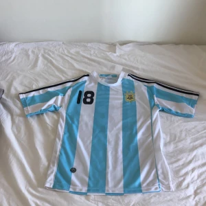 Argentina Messi 18 fotbollströja - Säljer en klassisk Argentina fotbollströja med Messi och nummer 18 på ryggen. Tröjan har blå och vita vertikala ränder, broderat AFA-emblem på bröstet och svarta detaljer på axlarna. Tillverkad i lätt och ventilerande material, perfekt för fotbollsfans. Och för andra som gillar Fotboll. Storlek M