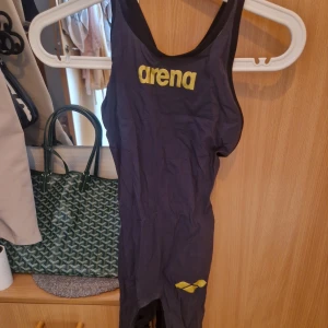 Mörkgrå baddräkt från Arena - Säljer en mörkblå baddräkt från Arena med guld logga framtill och på benet. Modellen har breda axelband och sportig passform som sitter tight mot kroppen. Perfekt för simträning eller tävling. Materialet är syntet och torkar snabbt. Stängd rygg, modell Air