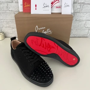 Svarta Christian Louboutin sneakers - Säljer ett par svarta sneakers från Christian Louboutin med ikonisk röd sula och svarta nitar på tån. Skorna har snörning och en rund tå, tillverkade i skinn och mocka för en lyxig känsla. Perfekt för dig som vill sticka ut med exklusiv design och edgy detaljer.