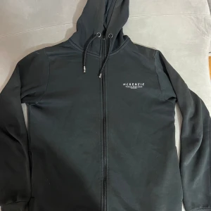 Svart McKenzie zipp  - Svart hoodie från McKenzie i storlek M med vit logga på bröstet. Tröjan har huva med snörning, hel dragkedja framtill och långa ärmar. 
