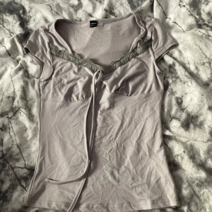 Ljuslila topp med spets från SHEIN - Supersöt ljuslila topp från SHEIN med korta ärmar, spetsdetalj vid urringningen och knytband framtill. Figurnära passform och mjukt material som känns skönt mot huden. Perfekt att styla med jeans eller kjol för en trendig look.