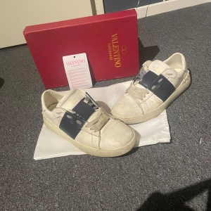 Valentino Garavani Open  - Snygga Valentino Garavani Open sneakers i vitt skinn med bred mörkblå rem över vristen och coola nitar baktill. Klassisk rund tå och platt sula. Kommer med originalkartong och dustbag – perfekt för dig som gillar exklusiva streetwear-vibbar.