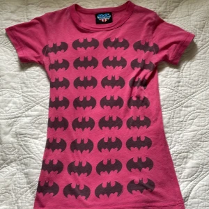 Rosa Batman t-shirt från Junk Food - Cool rosa t-shirt från Junk Food med svart Batman-logga i upprepade rader över hela framsidan. Tröjan är väl använd och är lite nopprig, och sömmen är sliten som man ser på bilden. Klassisk rund hals och korta ärmar. Materialet är mjuk bomull och passformen är normal. 