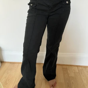Svarta bootcut byxor från H&M Divided - Svarta byxor från H&M Divided i bootcut-modell med hög midja. Byxorna har raka ben med lätt utsvängning och dekorativa fickor framtill med knappar. Tillverkade i bomullsmix med lite stretch för skön passform.