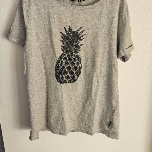 Grå t-shirt med paljettananas - Grå t-shirt från Pernilla Wahlgren i storlek S, med korta ärmar och rund hals. Framsidan har en stor ananas gjord av svarta och silvriga paljetter som ger en cool och glittrig vibe. Perfekt för dig som gillar att sticka ut med detaljer.