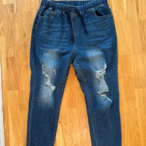 Blå jeansbyxor med resår och slitningar - Säljer ett par blå jeansbyxor med resår i midjan och snörning. Byxorna har slitna detaljer och hål på framsidan för en avslappnad streetstyle-look. Modellen är rak med normal passform och har fickor både fram och bak.