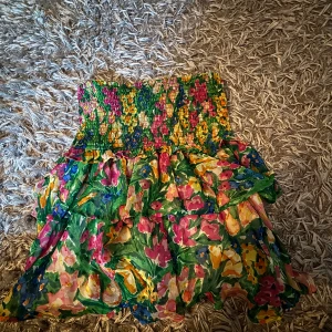 Blommig kjol från Lindex - Supersöt blommig kjol från Lindex i storlek S. Kjolen har smock upptill och volanger nedtill, med färgstarkt mönster i grönt, rosa, gult, blått och orange. Perfekt för dig som gillar färg och mönster! Materialet är lätt och luftigt, idealiskt för varma dagar.