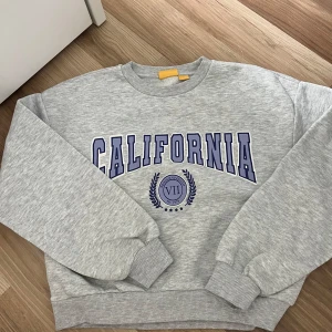 Grå sweatshirt CALIFORNIA Gina Tricot - Grå sweatshirt från Gina Tricot med tryck i blått och vitt där det står CALIFORNIA och ett emblem framtill. Tröjan har rund hals, ribbade muddar och en loose passform. Perfekt för en chill och sportig look.