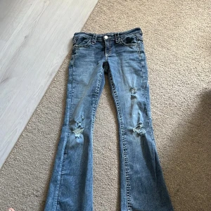Blå bootcut lågmidjade jeans med slitningar - Säljer ett par blå bootcut jeans med snygga slitningar på båda benen. Jeansen har klassisk femficksdesign, låg midja och är tillverkade i mjukt denimtyg. Perfekta för dig som gillar en avslappnad och trendig look.