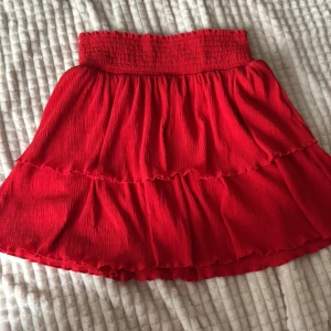 Röd volangkjol från Kappahl - Supersöt röd kjol från Kappahl med volanger och smock i midjan. Kjolen har en kort och flowy passform med plisserat tyg som ger en snygg struktur. Perfekt för dig som gillar färgstarka och trendiga plagg.