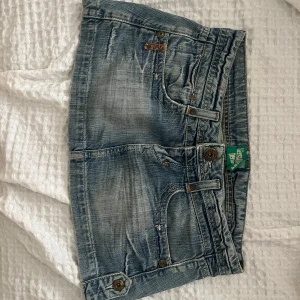 Blå jeanskjol  - Säljer en kort jeanskjol  i ljusblå tvätt med slitningar och coola detaljer. Kjolen har klassiska fickor fram och bak, bälteshällor och snygga knappar. På baksidan finns en patch med stjärnor och paljetter för extra edge.
