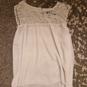 Vit blus med spets från Zanzea - Supersöt vit blus från Zanzea med vacker virkad spets upptill och ärmlös design. Blusen har en luftig, flowy passform och dubbla lager tyg nedtill för extra snygg effekt. Perfekt att matcha med jeans eller kjol för en fräsch look.