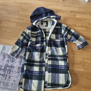 Rutig overshirtjacka från Zara - Säljer en oversized rutig overshirtjacka från Zara i storlek S. Jackan har svart, beige, grå, lila och gröna toner, knappar framtill och två bröstfickor. Materialet är mjukt och den har en avtagbar marinblå huva. Perfekt för lager-på-lager-stil.
