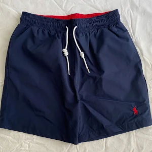 Marinblå badshorts från Polo Ralph Lauren - Snygga marinblå badshorts från Polo Ralph Lauren med röd insida i midjan och vit snörning framtill. Klassisk Polo-logga broderad i rött på benet. Shortsen har elastisk midja och en bakficka. Använda enbart en gång. Storlek M men passar mer för S. Priset kan diskuteras 😊