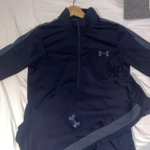 Under Armour Tracksuit - Snyggt mörkblått träningsset från Under Armour med jacka och matchande byxor. Jackan har dragkedja framtill och logga på bröstet, medan byxorna har logga på benet och diskreta sidopaneler. Perfekt för gymmet eller löprundan.