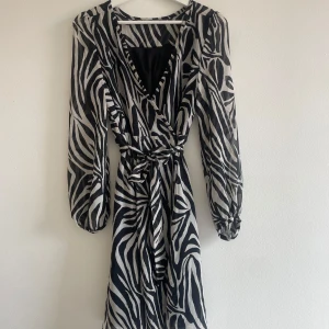 Zebramönstrad omlottklänning  - Snygg kort omlottklänning från Zara i svart och vitt zebramönster. Klänningen har långärmade transparenta ärmar, v-ringning och knytband i midjan. Materialet är lätt och luftigt, perfekt för att sticka ut med coola vibes. Passformen är normal och modellen är feminin. Använd 1 gång 