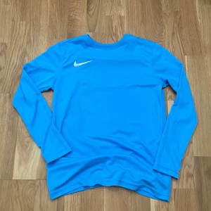 Blå långärmad Nike tröja - Snygg blå långärmad tröja från Nike med klassisk vit swoosh på bröstet. Tröjan har rund hals och är gjord i ett mjukt och lätt polyestermaterial som andas. Perfekt för träning eller chill dagar. Enkel design med sportig vibe.