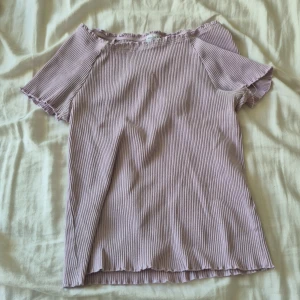 Ljuslila ribbad topp från Lindex - Supersöt ljuslila ribbad topp från Lindex med korta ärmar och offshoulder-modell. Toppen har volangkant vid halsringning och nederkant för en extra gullig touch. Perfekt för dig som gillar en enkel men ändå trendig stil.
