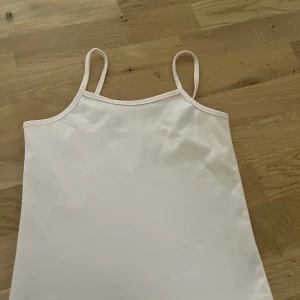 Vit linnetopp med smala axelband - Enkel och stilren vit linnetopp med smala axelband. Toppen har en rak passform och är tillverkad i ett mjukt, lätt material som känns skönt mot huden. Perfekt att matcha med jeans eller shorts för en clean look.