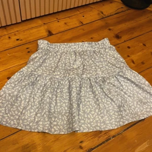 Blommig ljusblå volangkjol - Supersöt ljusblå kortkjol med vita små blommor och volangdetaljer. Kjolen har en mjuk resår i midjan och är gjord i ett lätt bomullstyg som känns skönt mot huden. Perfekt för dig som gillar romantiska och somriga vibes.