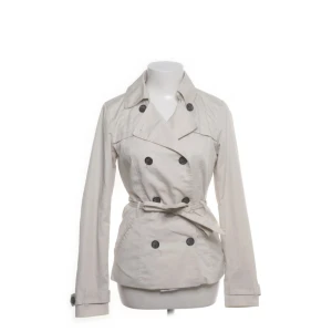 Beige trenchcoat - Snygg beige trenchcoat med dubbelknäppning och bälte i midjan. Jackan har klassisk krage, långa ärmar och svarta knappar framtill samt vid ärmsluten. Perfekt för dig som vill ha en stilren och tidlös look.