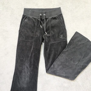 Svarta Juicy Couture mjukisbyxor - Svarta mjukisbyxor från Juicy Couture i mjuk velour med bred resår och snörning i midjan. Byxorna har stora fickor fram och bakfickor med silverfärgade knappar. Broderad JC-logga framtill ger en lyxig touch. Byxorna är också uppsydda lite.