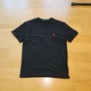 Svart t-shirt från Polo Ralph Lauren - Svart t-shirt från Polo Ralph Lauren med klassisk rund hals och korta ärmar. På bröstet finns den ikoniska röda Polo-loggan broderad. T-shirten är gjord i mjuk bomull och har en clean, stilren look som funkar till jeans eller shorts.