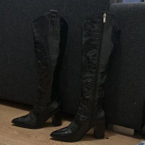 Svarta stövlar med klack - Snygga svarta boots med högt skaft och krokodilpräglat mönster. Skorna har spetsig tå, kraftig blockklack och dragkedja på insidan. Tillverkade i glansigt syntetmaterial som ger en edgy och trendig look. Aldrig användna utomhus 